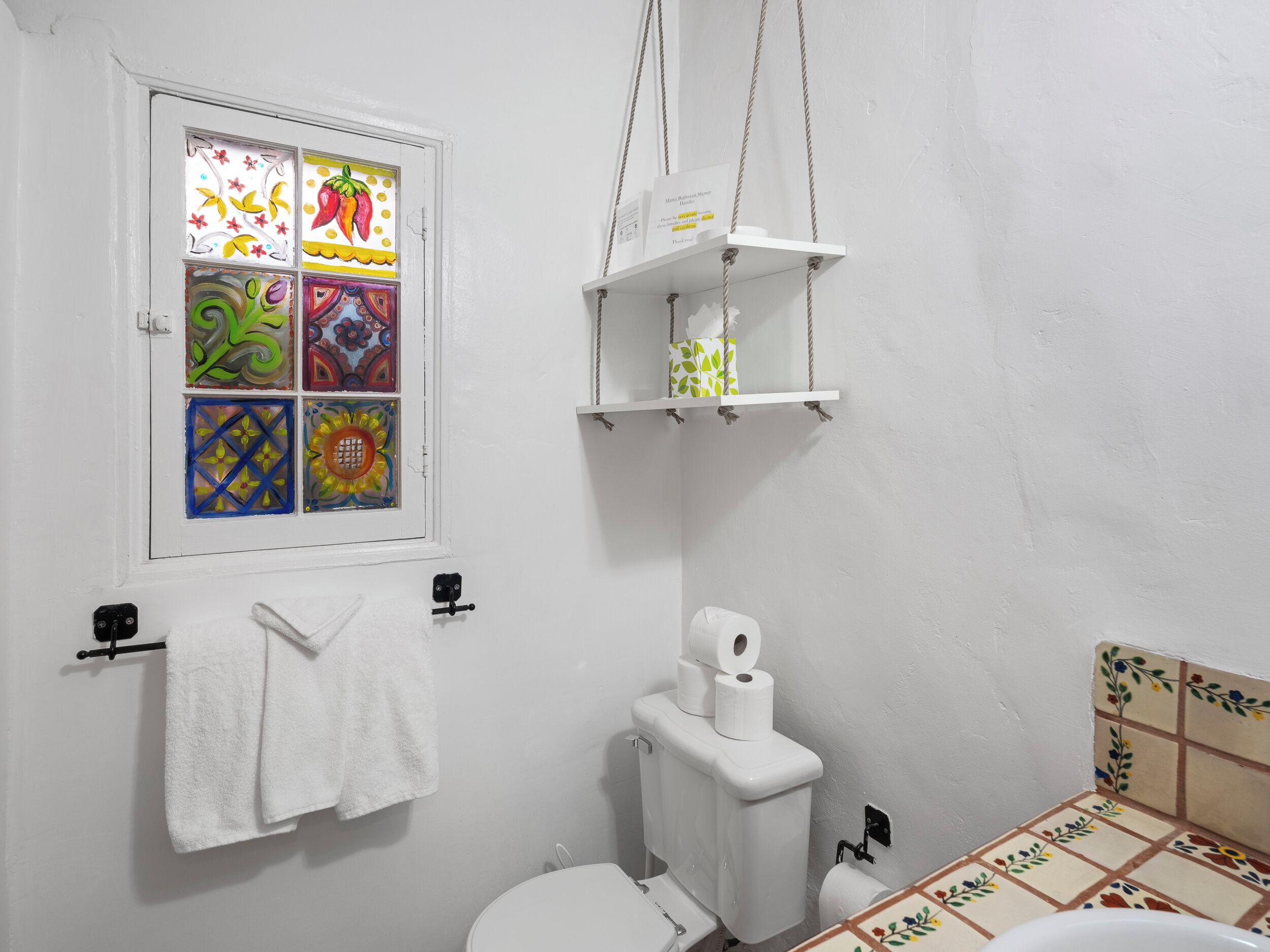 Une salle de bain avec des toilettes blanches, une fenêtre ornée de vitraux colorés et une étagère suspendue où sont rangés les articles de toilette.