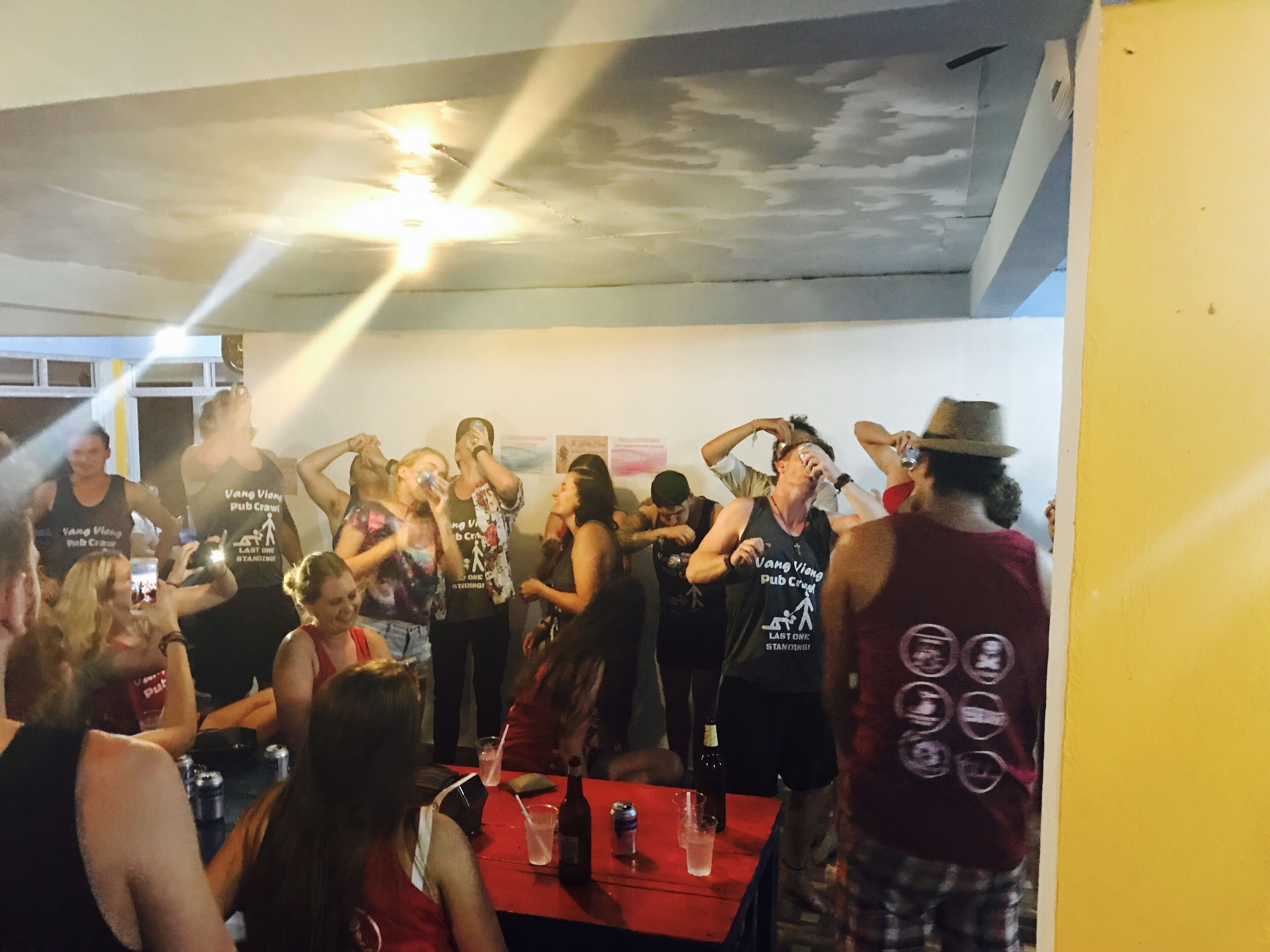 A group of people in a room with a red table, some wearing black tank tops with text.
