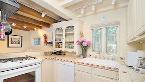 A kitchen with white cabinets, a stove, and a dishwasher. There is a window with a view of trees and a clock on the wall. - Casa Archuleta | Charming Historic (Santa Fe)