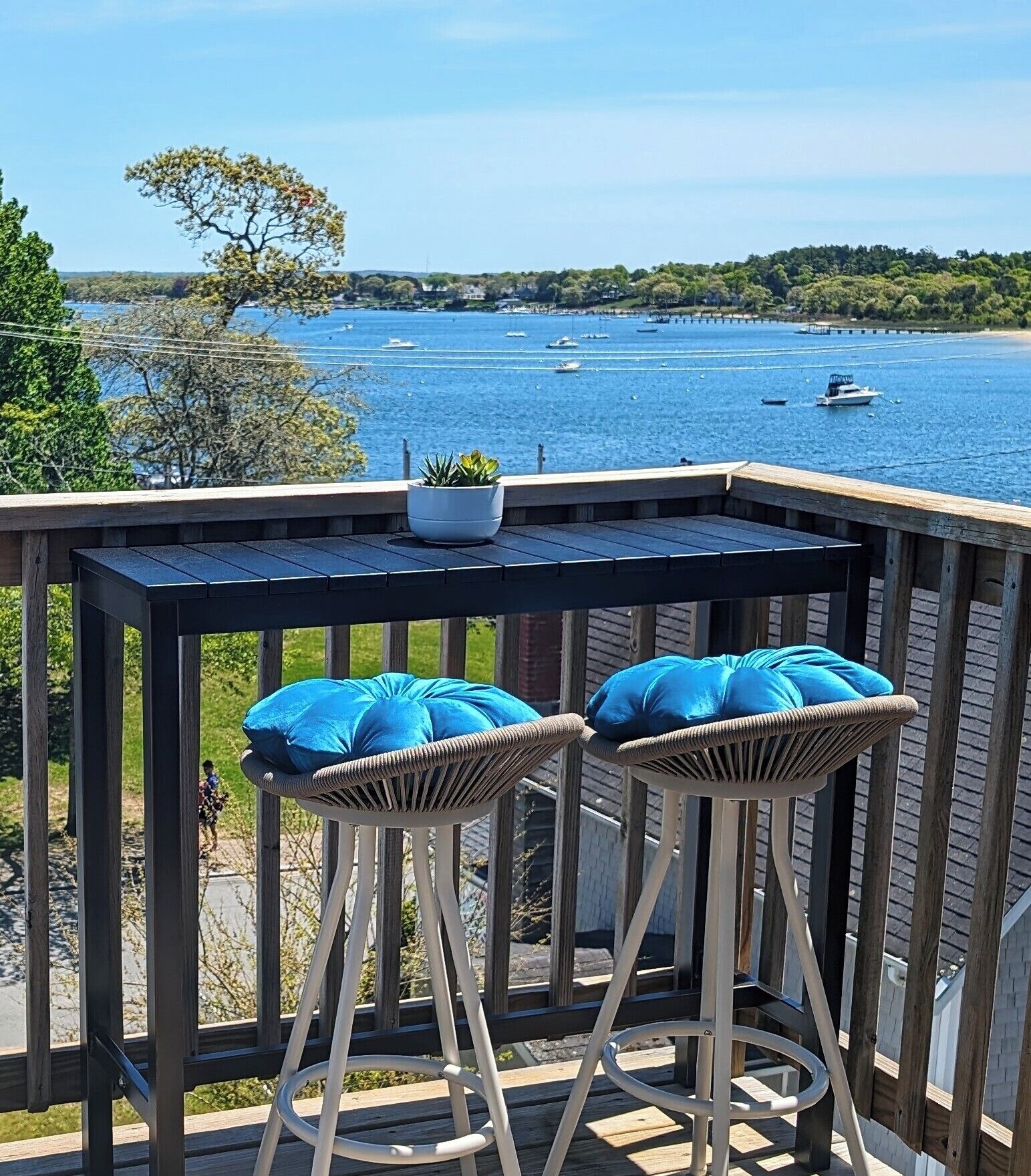 Una terraza de madera con dos sillas de mimbre y una mesa, con vista a un cuerpo de agua con barcos.