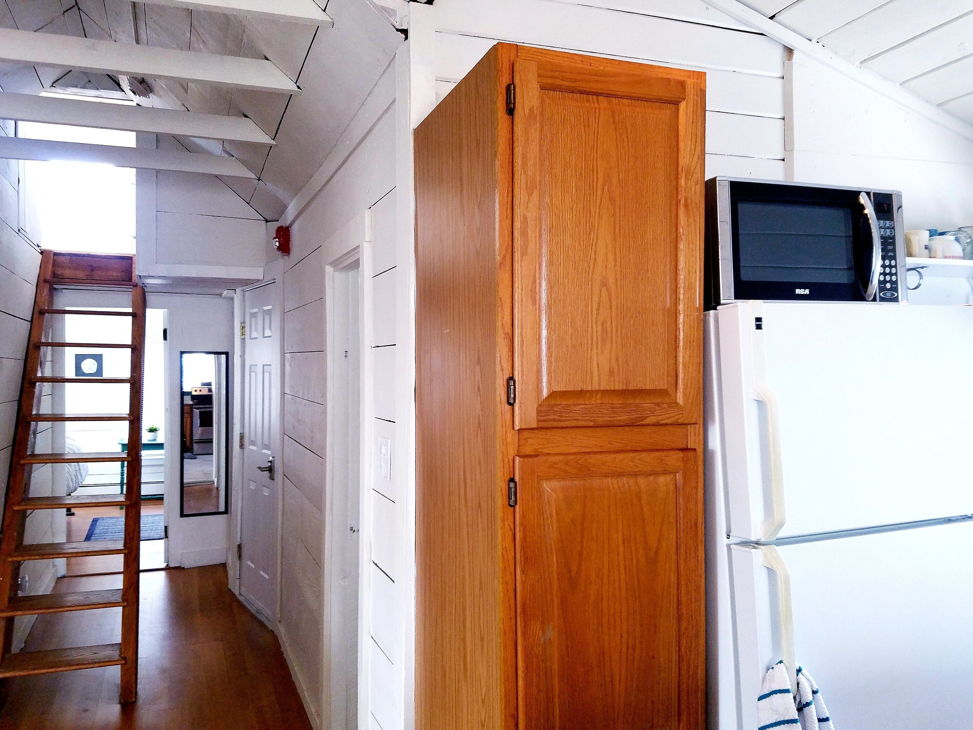A kitchen with white cabinets, a wooden door, a microwave, and a refrigerator. A wooden ladder is leaning against the wall.