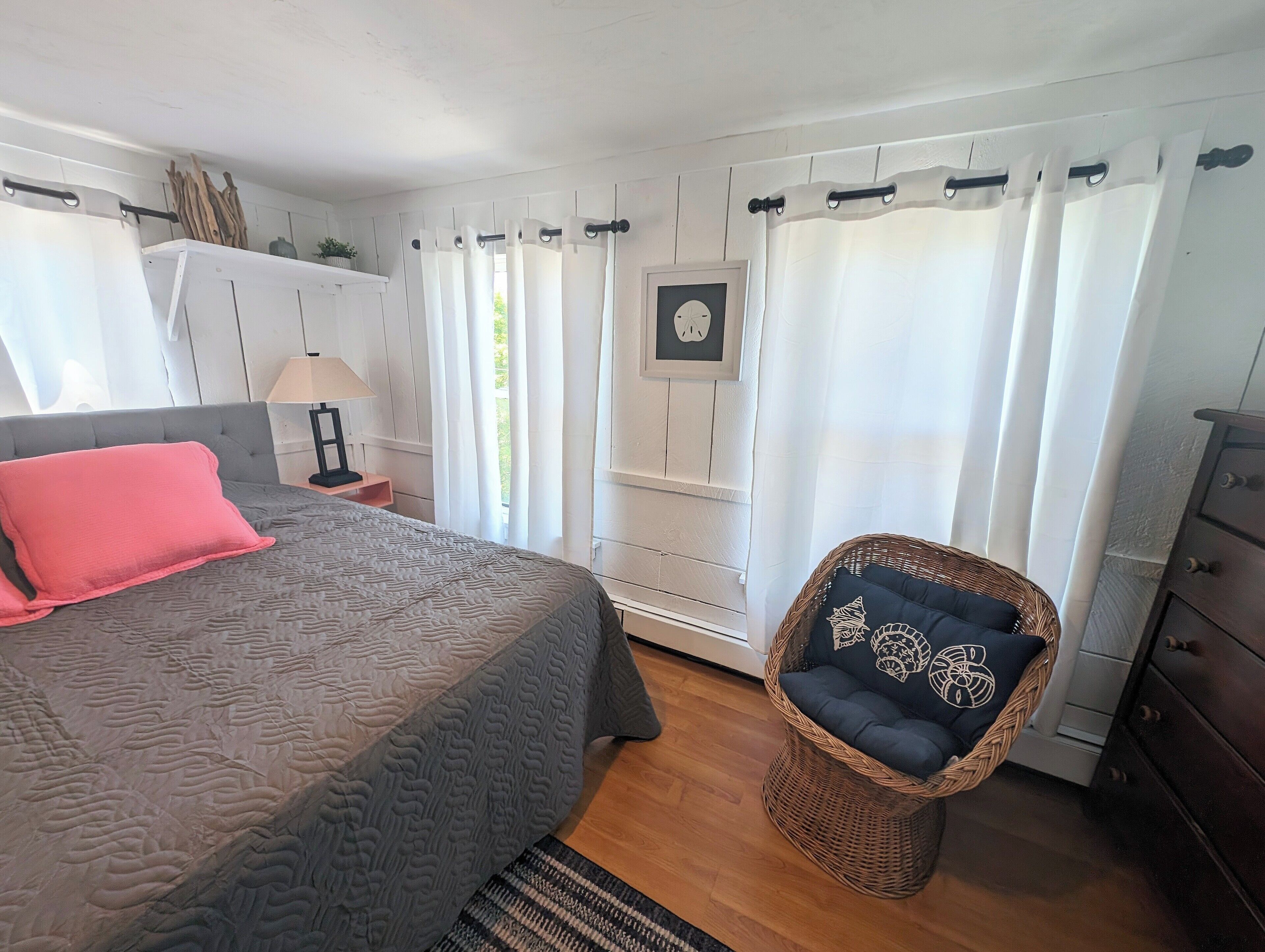 A bedroom with a bed, a nightstand, a lamp, a wicker chair, and a dresser.