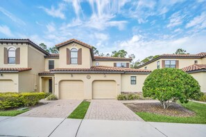 A residential building with a beige facade, brown shutters, and a tiled roof. There are two garage doors, a small tree, and a well-maintained lawn. - Charming Townhome | Gated Resort with Pool (Davenport)