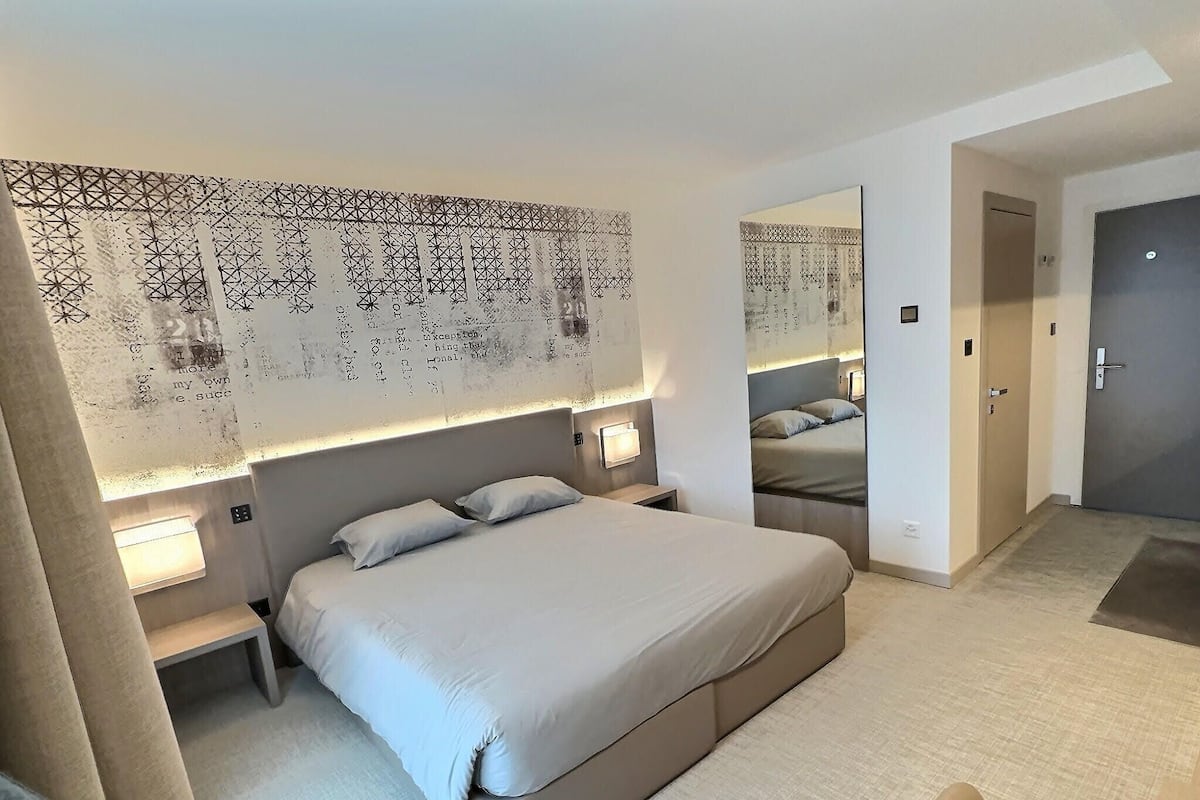 Habitación de hotel moderna con una cama grande, mesitas de noche, un espejo y un papel tapiz estampado.