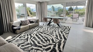 A modern living room with a sofa, a dining table with chairs, and a large window offering a view of the outdoors.