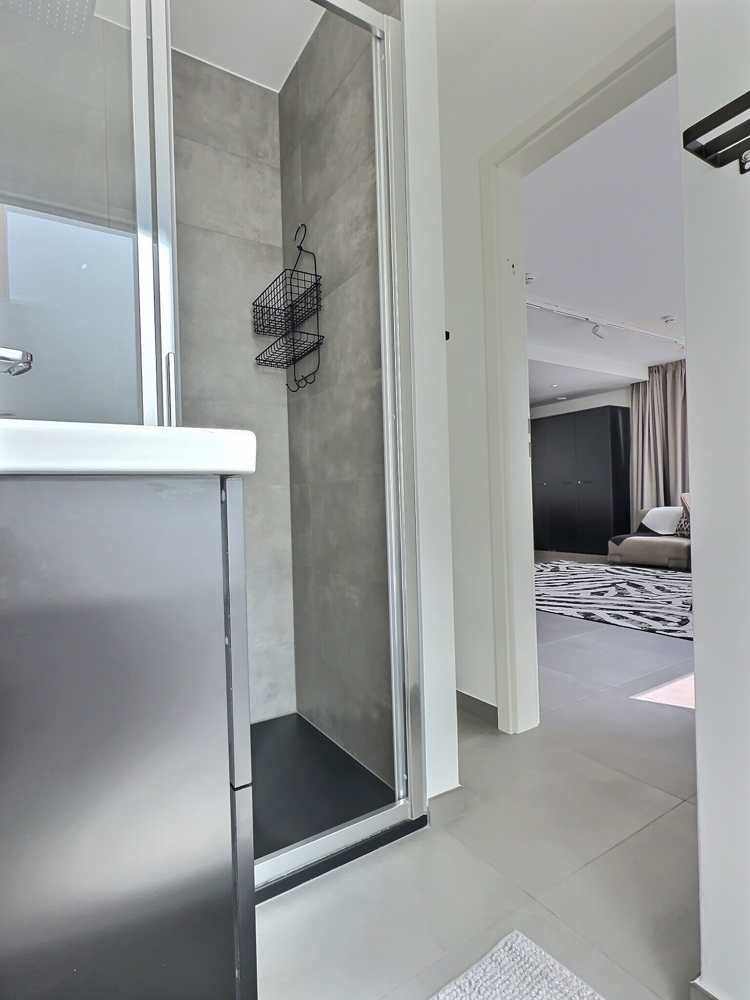 A modern bathroom with a glass shower enclosure, a white sink, and a black wire basket on the wall.