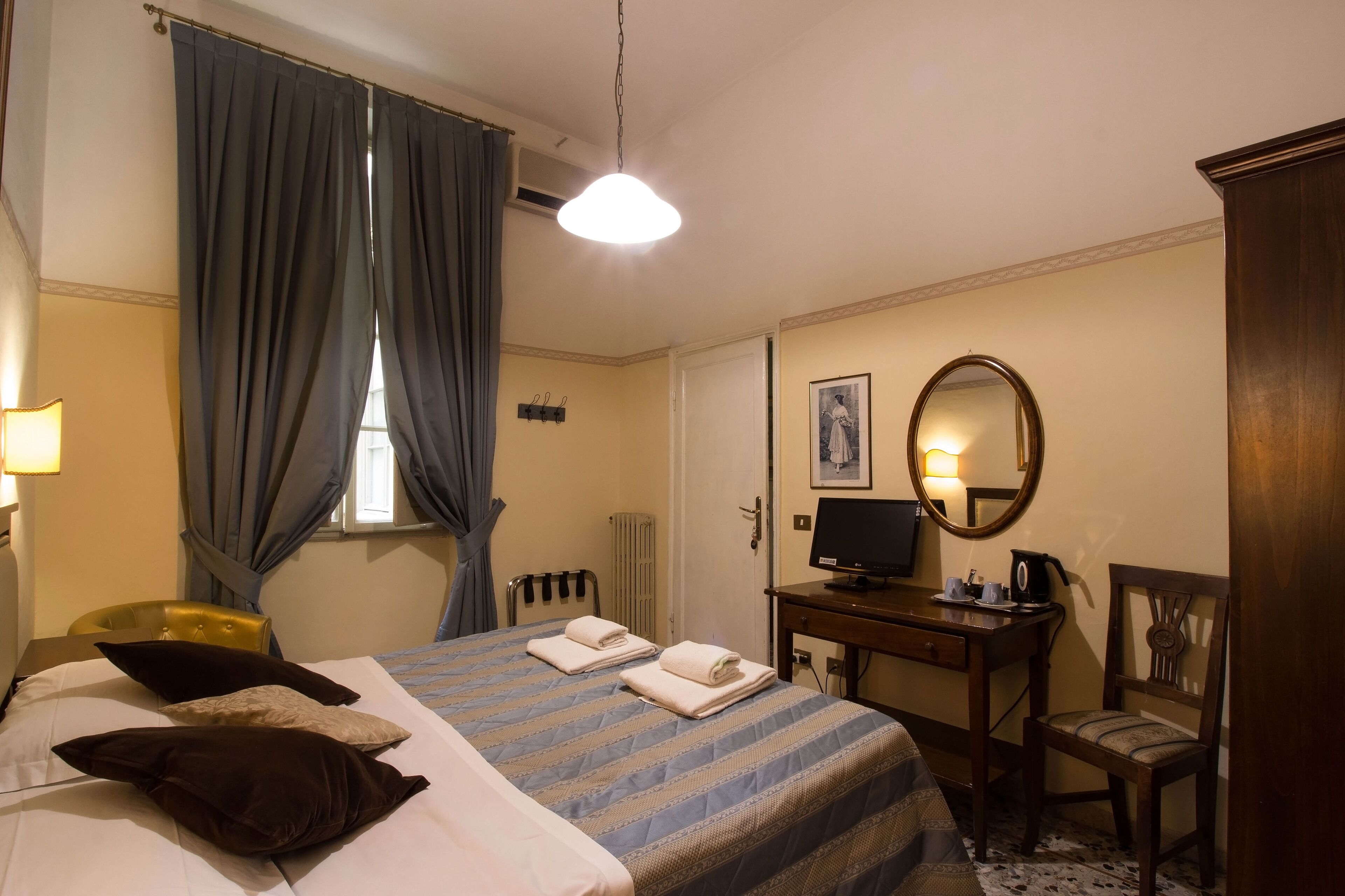 A hotel room with a bed, a desk with a computer, a chair, a mirror, and a window with curtains.