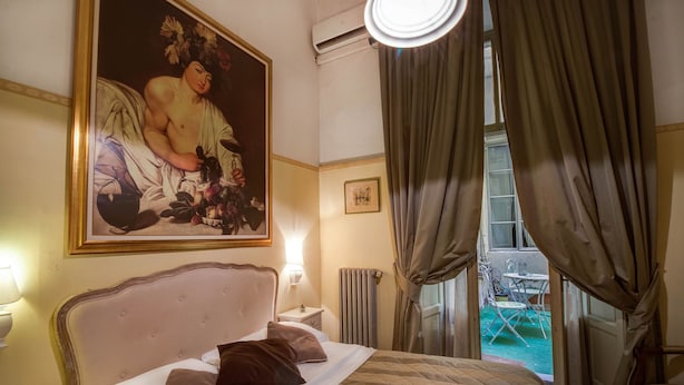 A hotel room with a large bed, a nightstand, a painting, a window with curtains, and a view of the outside. - Tourist House Duomo (Florence)