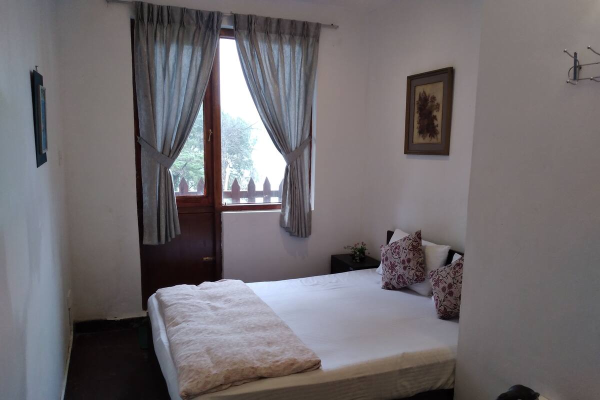 Une chambre à coucher avec un lit, une fenêtre avec des rideaux, une table de chevet avec une bouilloire et un tableau encadré au mur.