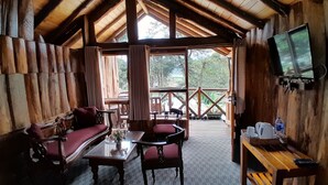 A wooden cabin with a TV mounted on the wall, a sofa, chairs, a table with a vase of flowers, and a wooden deck outside. - Eco Creations Nuwaraeliya (Nuwara Eliya)