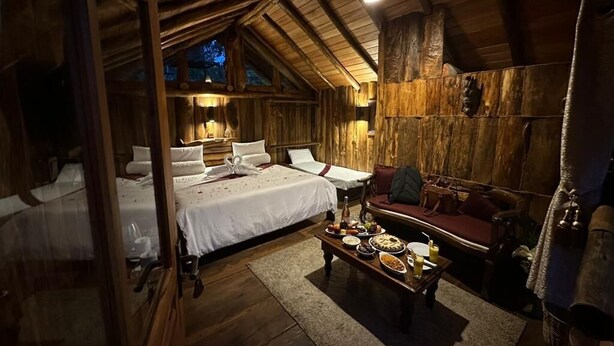 A rustic wooden cabin with a cozy interior, featuring a bed, a sofa, and a dining table set with food. - Eco Creations Nuwaraeliya (Nuwara Eliya)