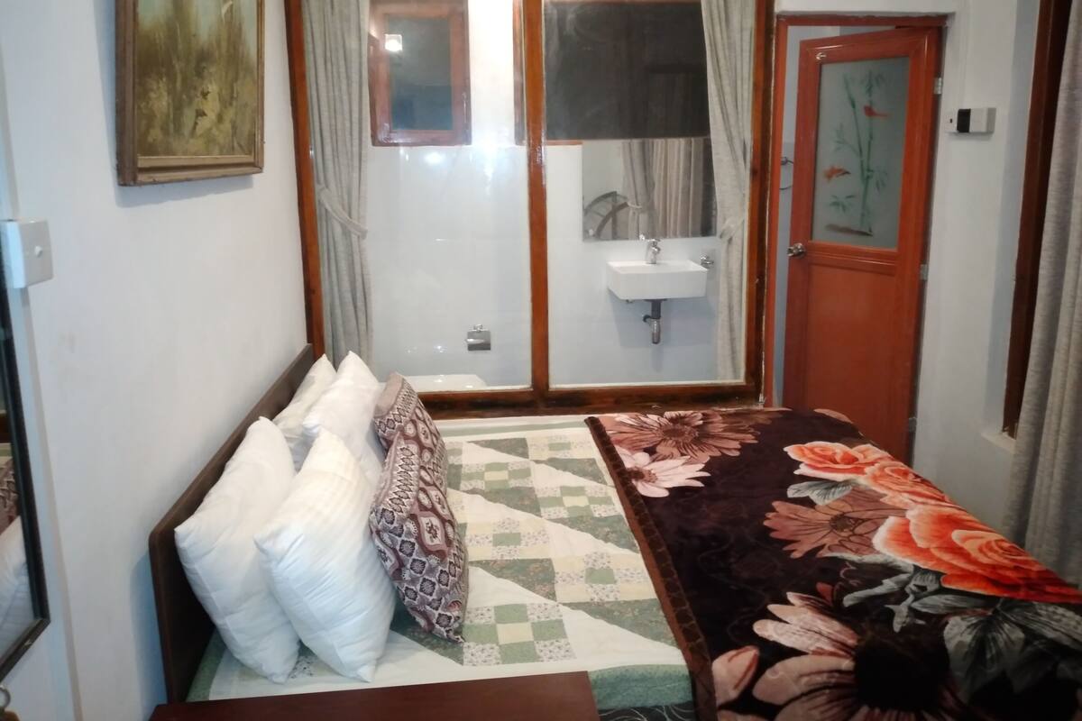 Une chambre à coucher comprenant un lit, une table de chevet, un miroir, une porte et une salle de bain visible à travers une porte en verre.