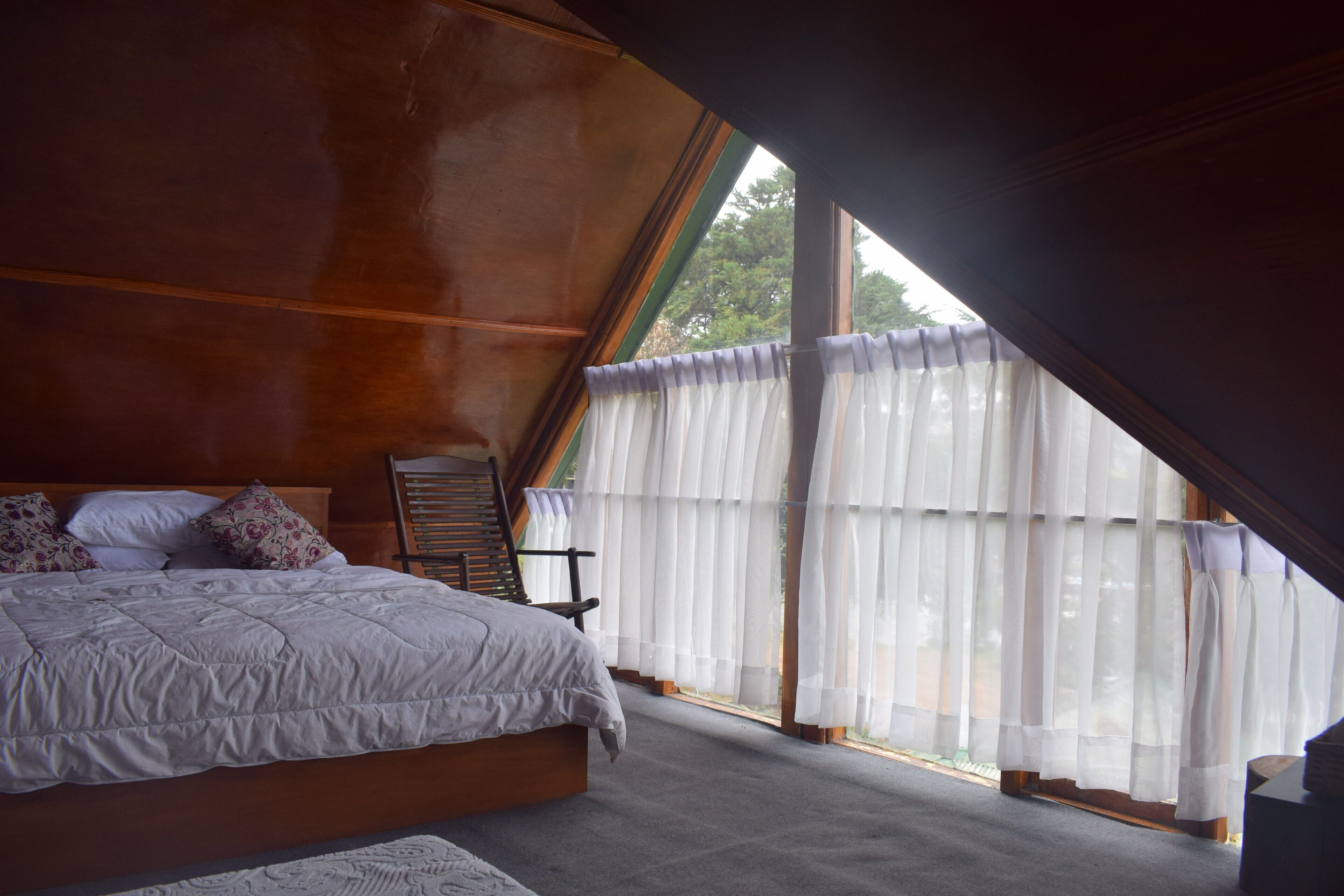 Un dormitorio con una cama grande, cabecera de madera y vistas al exterior a través de cortinas translúcidas.