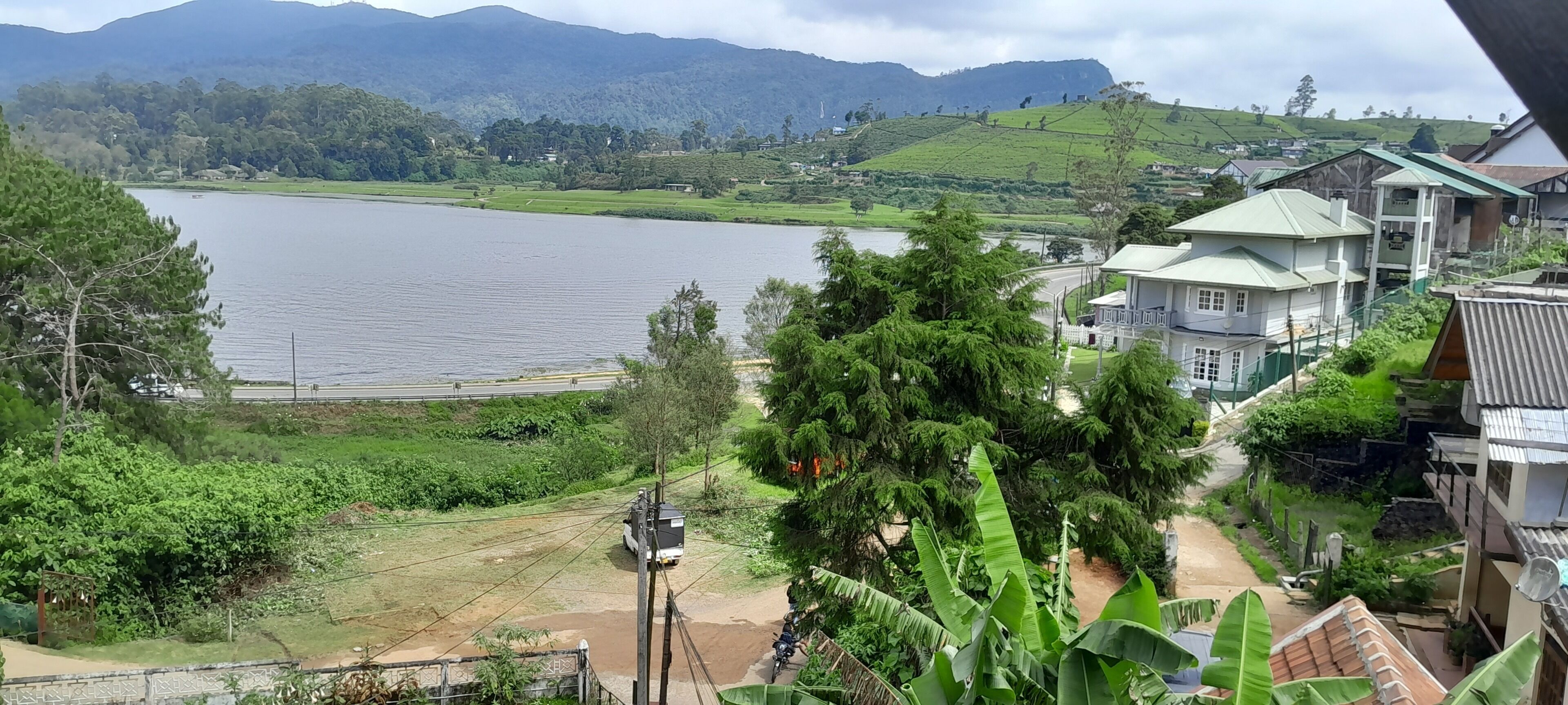 Vista de un lago, con una carretera en primer plano y una zona residencial con casas al fondo.
