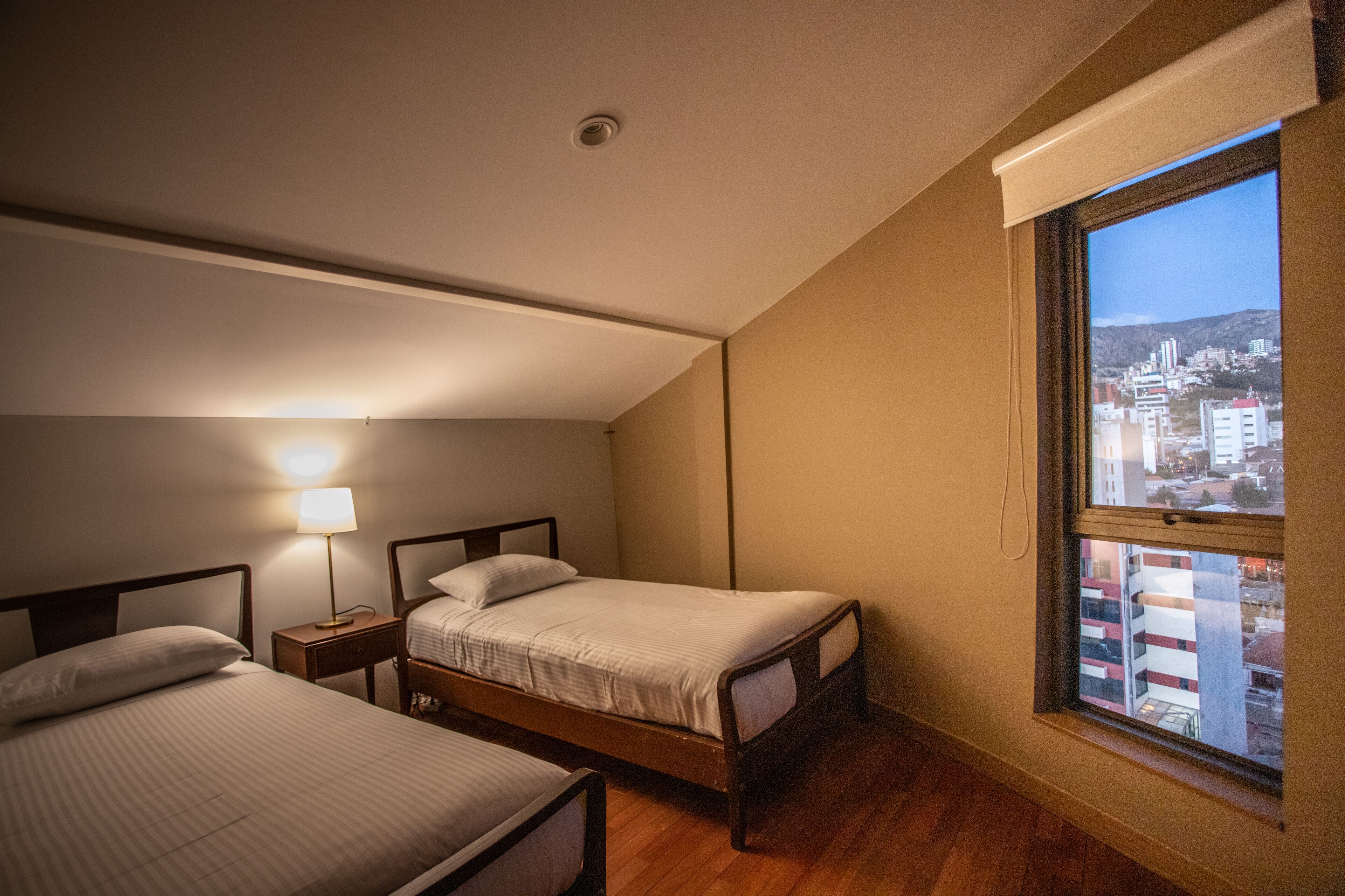 A bedroom with two beds, a nightstand with a lamp, and a window with a city view.