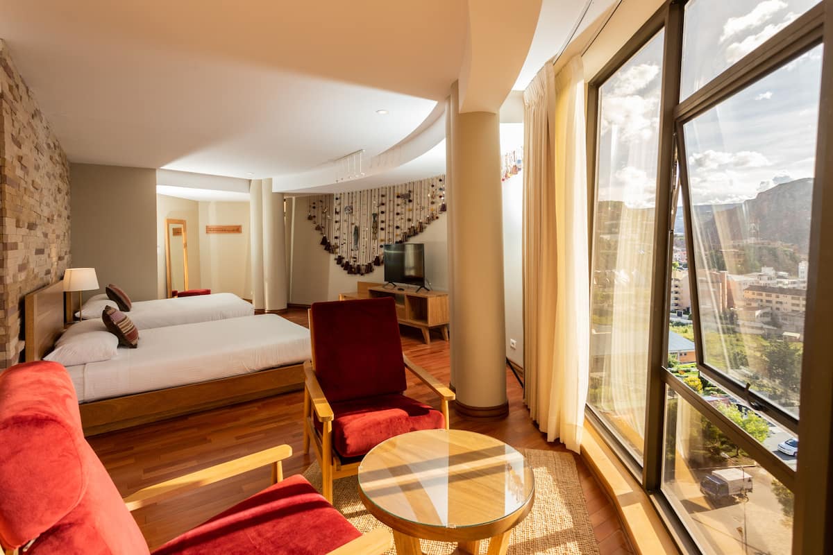 Habitación de hotel con una cama grande, un sillón rojo, una mesa de centro de vidrio y vistas a la ciudad a través de amplios ventanales.