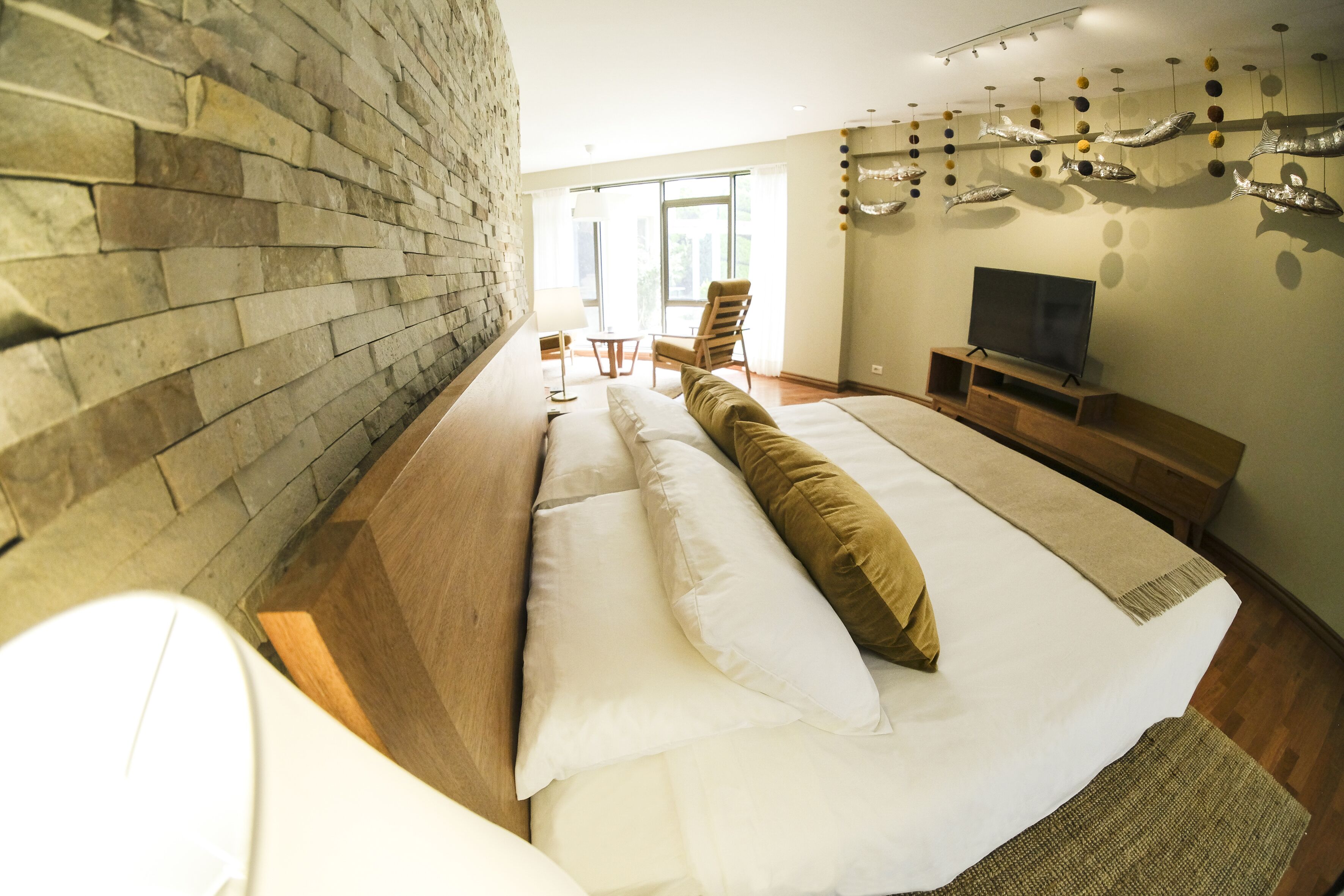 Una habitación de hotel moderna con pared de piedra, una cama grande con ropa de cama blanca y marrón, un televisor de pantalla plana, una mesita de noche de madera y un gran ventanal con vistas a zonas verdes.