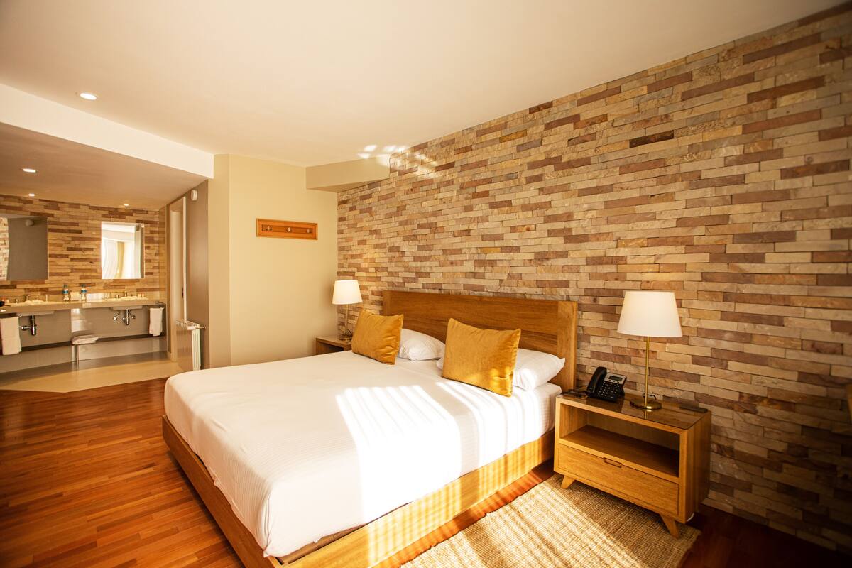 Habitación de hotel con una cama grande, mesitas de noche de madera, pared con diseño de ladrillo y baño con tocador y espejo.
