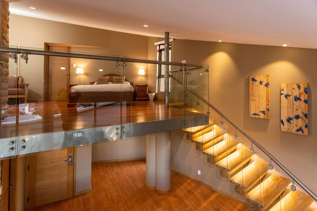 Una habitación de hotel moderna con balcón de vidrio, suelo de madera, una cama, una mesita de noche y un panel de madera en la pared.