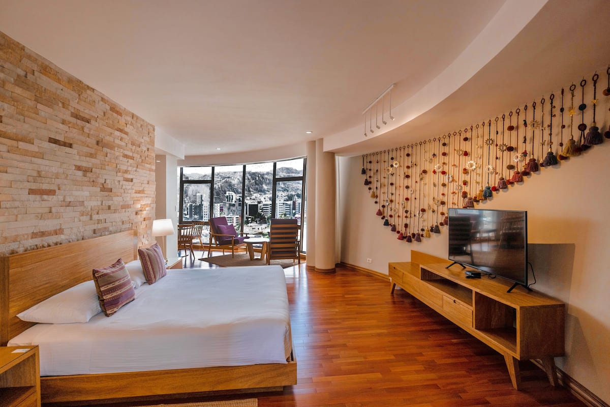 Una habitación de hotel moderna con una cama grande, un televisor, un mueble de televisión de madera, una pared de ladrillo y vistas de la ciudad a través de amplias ventanas.