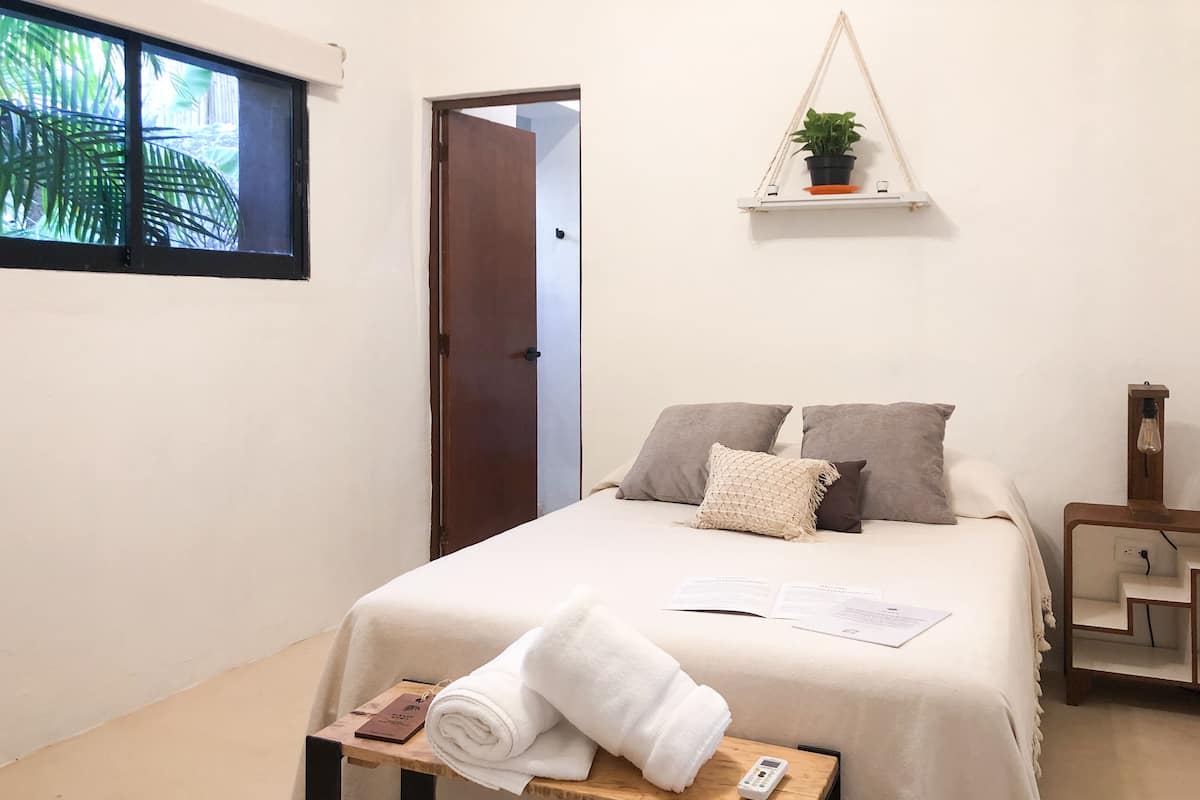 A neatly made bed with pillows, a bedside table with a towel, a small shelf with a plant, and a door leading to another room.