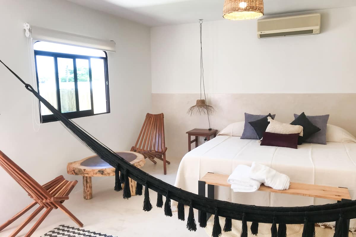 A bedroom with a large bed, a wooden chair, a small table, a hanging light, and a window with blinds.