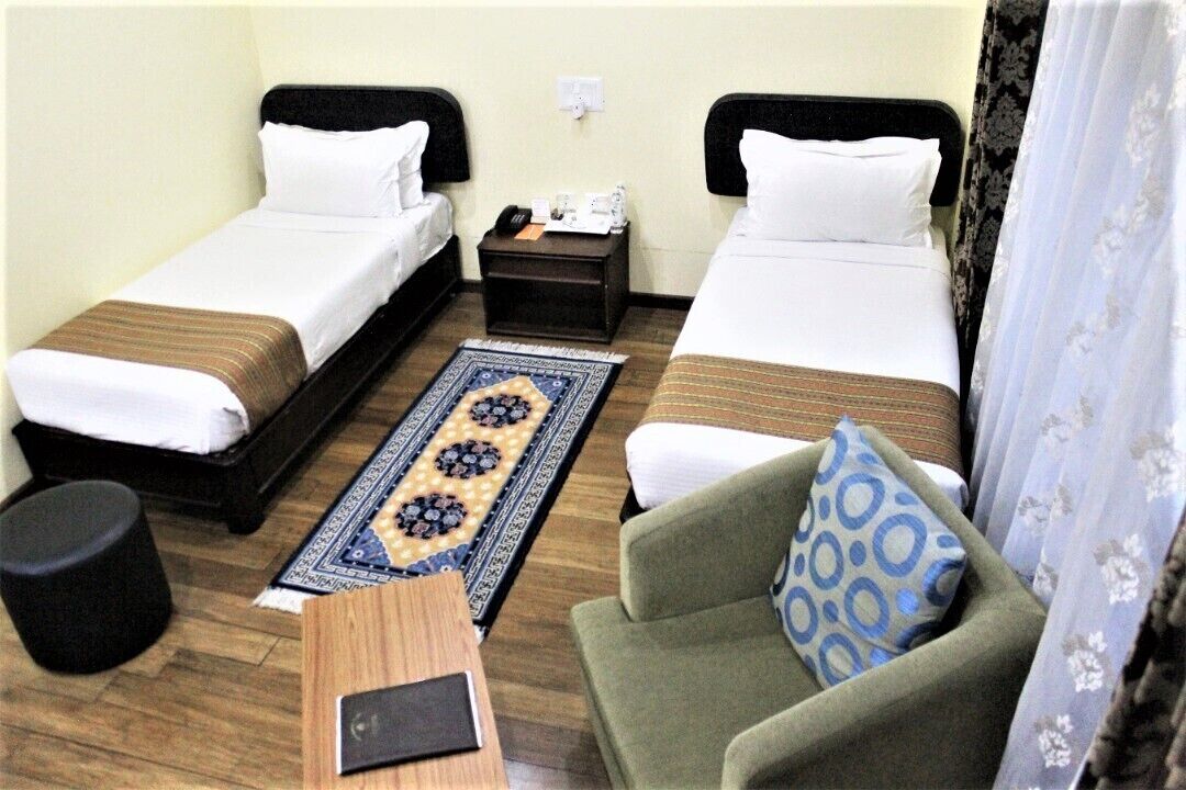 A hotel room with two single beds, a small table, a chair, and a patterned rug.