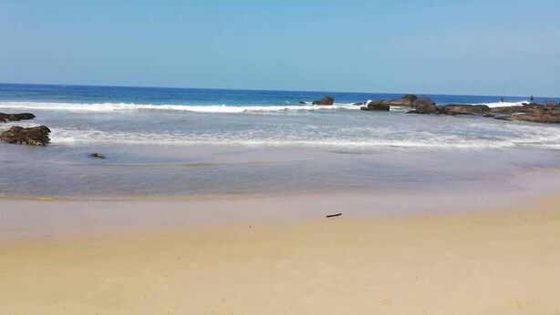 Una playa con cielos despejados, olas tranquilas y afloramientos rocosos.