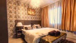 Une chambre avec un papier peint à motifs, un lit recouvert d’une couette blanche, une table de chevet, une lampe et un banc avec un revêtement à motifs.