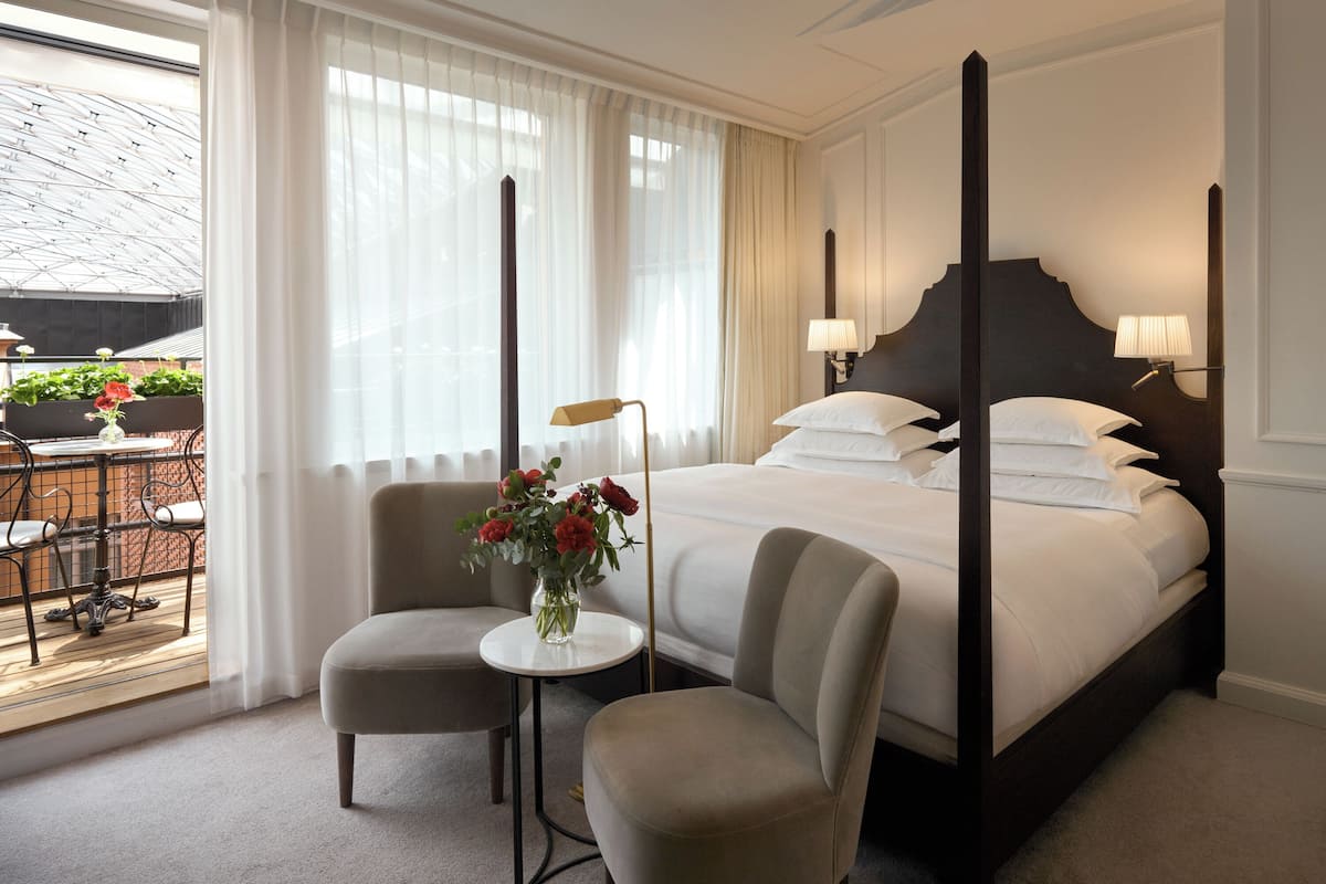 A four-poster bed with a dark headboard, two armchairs, a small table with a vase of flowers, and a view of the outdoors through large windows.