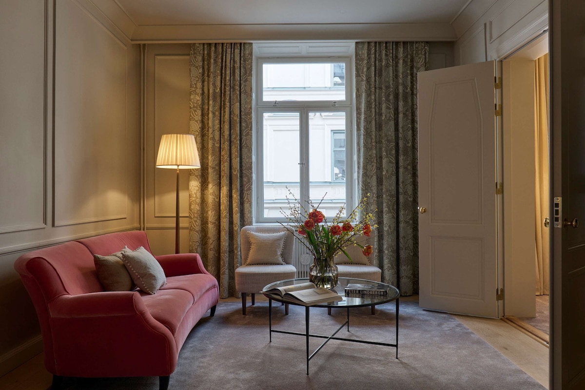 A living room with a red sofa, a white armchair, a glass coffee table with a book and flowers, and a lamp.