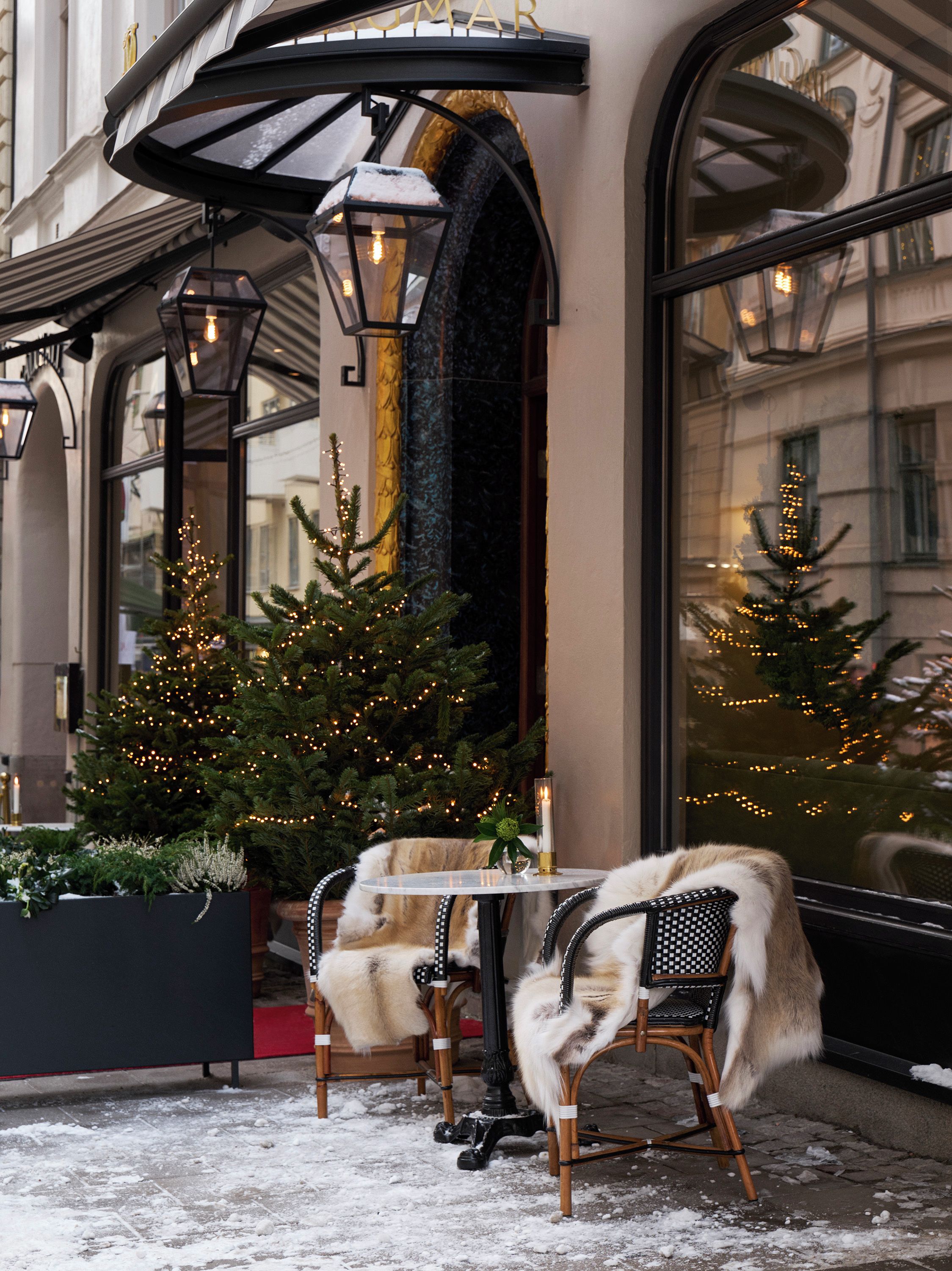 A snow-covered outdoor seating area with two chairs and a table, a Christmas tree with lights, and a building facade with large windows and a sign.