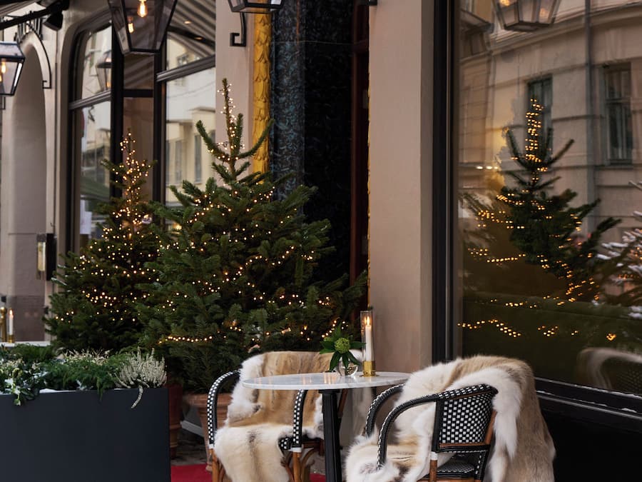 A snow-covered outdoor seating area with two chairs and a table, a Christmas tree with lights, and a building facade with large windows and a sign.