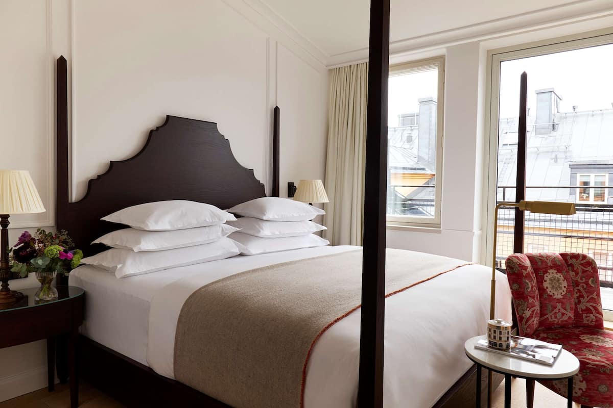 A hotel room with a large bed, a small table with a lamp, a chair, and a view of the city through the window.
