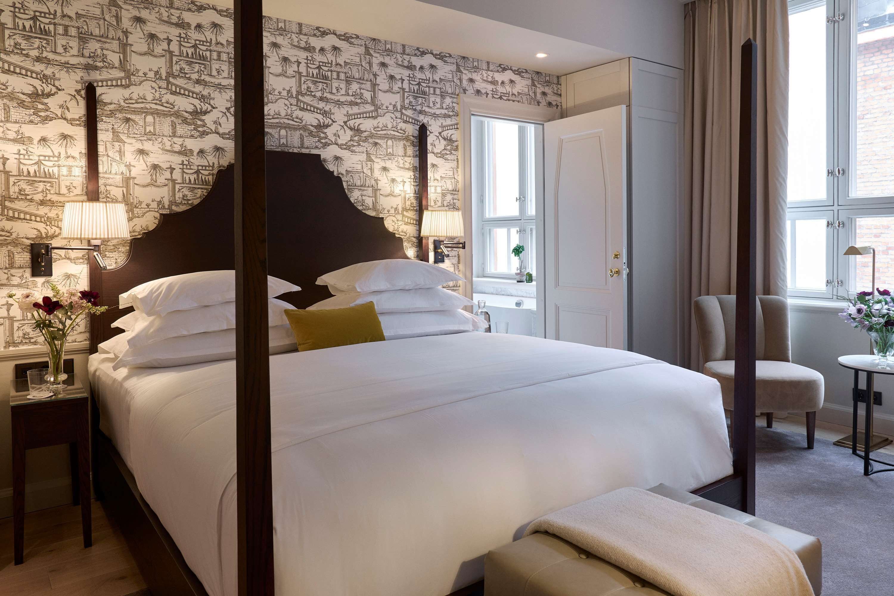 Una camera d'albergo con un letto grande, un comodino con una lampada, una sedia e una finestra con tende.
