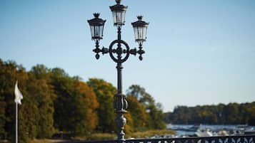 Een ijzeren straatlamp met vier lichten en een decoratieve knop, voor een heldere lucht en een waterpartij met boten.