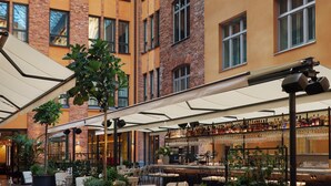 A spacious indoor courtyard with multiple dining tables and chairs, surrounded by brick and yellow buildings, and a glass ceiling above. - Villa Dagmar (Stockholm)