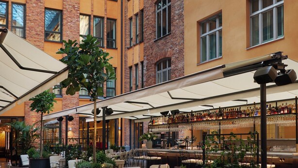 A spacious indoor courtyard with multiple dining tables and chairs, surrounded by brick and yellow buildings, and a glass ceiling above. - Villa Dagmar (Stockholm)