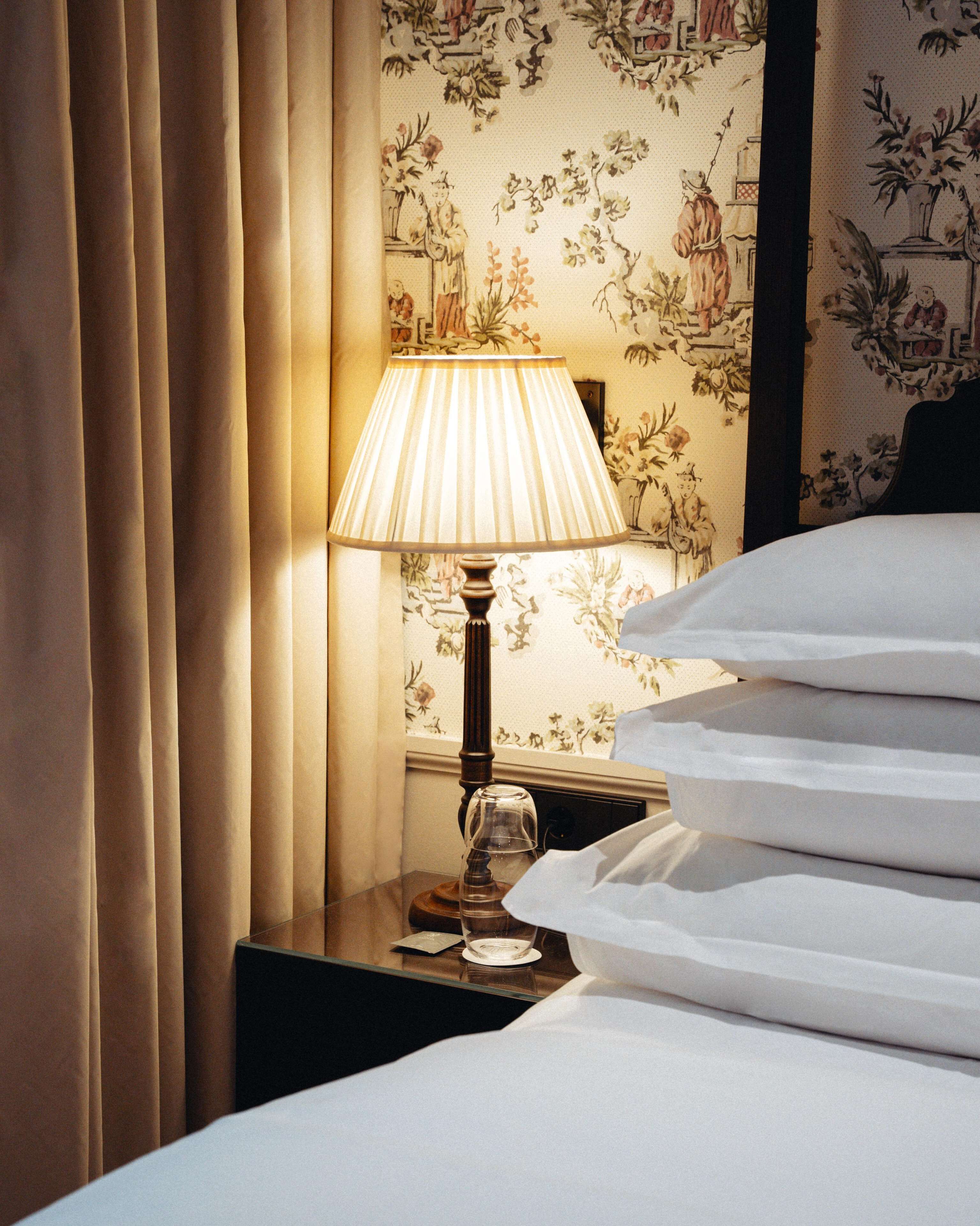 Una cama bien hecha con sábanas blancas, una lámpara de noche y un papel tapiz floral de fondo.