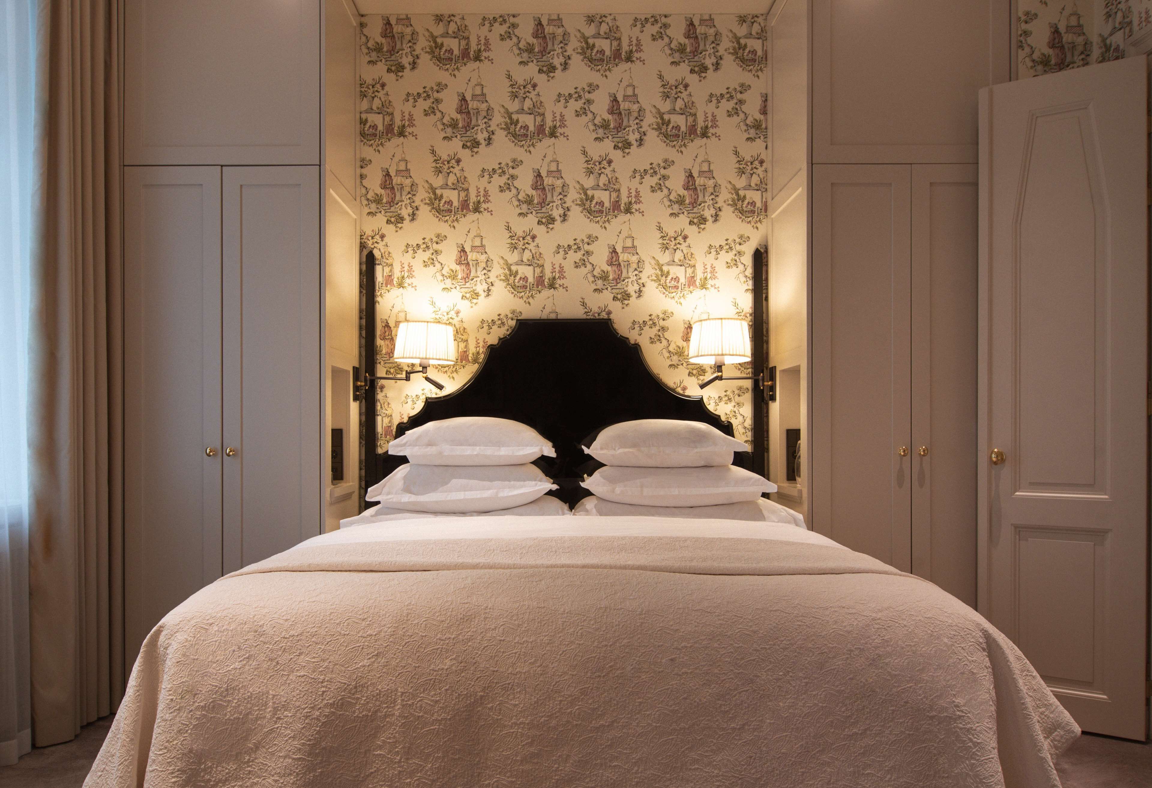 Una cama bien tendida con un cabecero negro, dos lámparas de pared y un estampado floral en el papel tapiz.