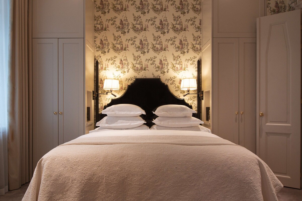 A neatly made bed with a black headboard, two wall-mounted lamps, and a floral wallpaper pattern.