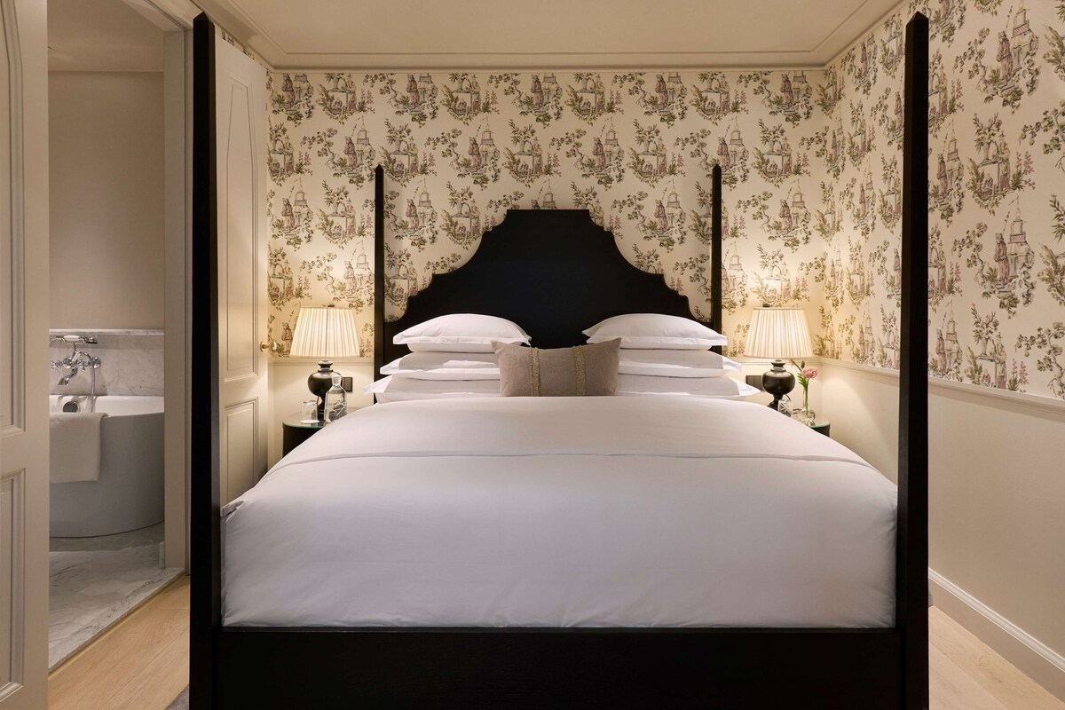 A four-poster bed with white linens, two bedside tables with lamps, and a bathroom with a bathtub and sink.