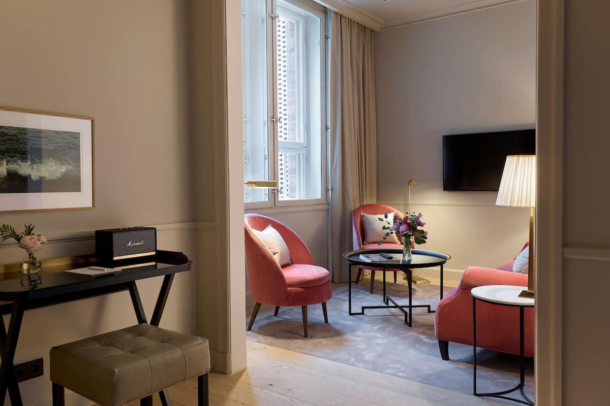 A modern hotel room with a desk, two pink armchairs, a small table with a vase of flowers, a flat-screen TV mounted on the wall, and a window with curtains.