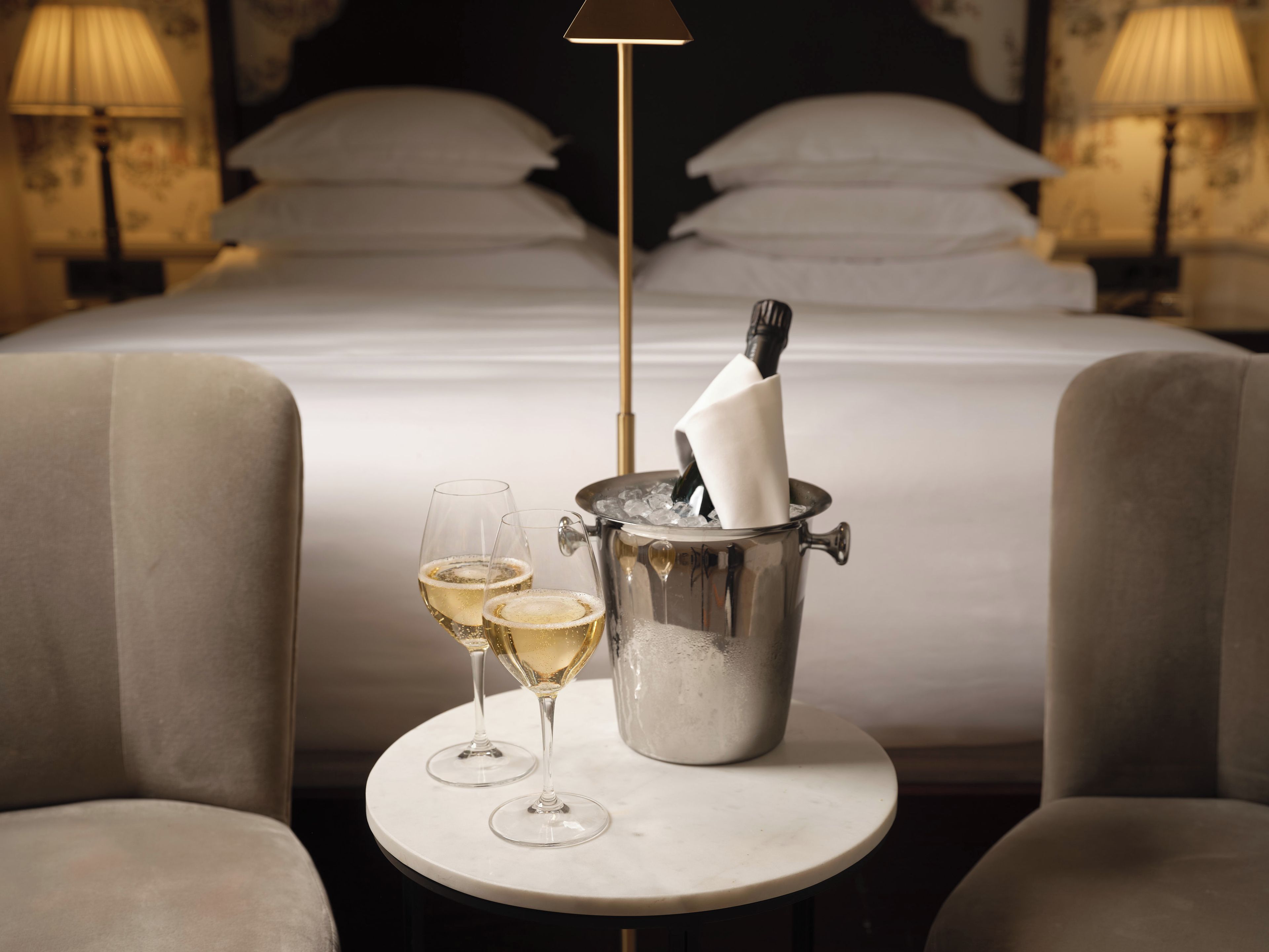 Een bed met witte lakens, een nachtkastje met twee champagneglazen, en een champagnekoeler met een fles en een servet.