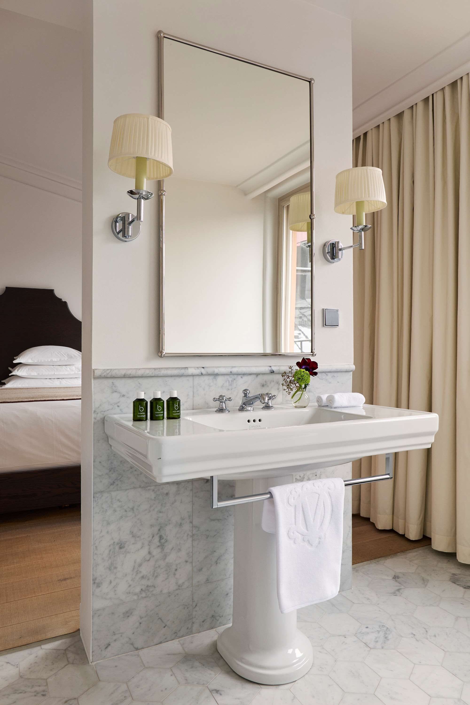 Un baño de hotel con un lavabo blanco, dos lámparas fijadas a la pared, un espejo grande y una cama en la habitación contigua.