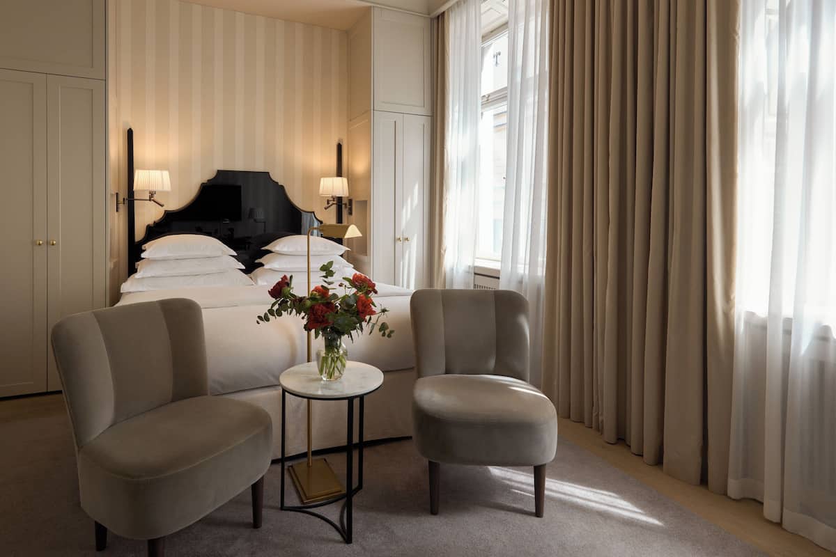 A hotel room with a bed, two armchairs, a small table with a vase of flowers, and a large window with curtains.