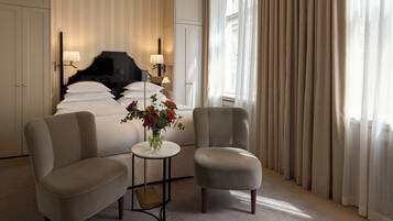 Ein Hotelzimmer mit einem Bett, zwei Sesseln, einem kleinen Tisch mit Blumenvase und einem großen Fenster mit Vorhängen.