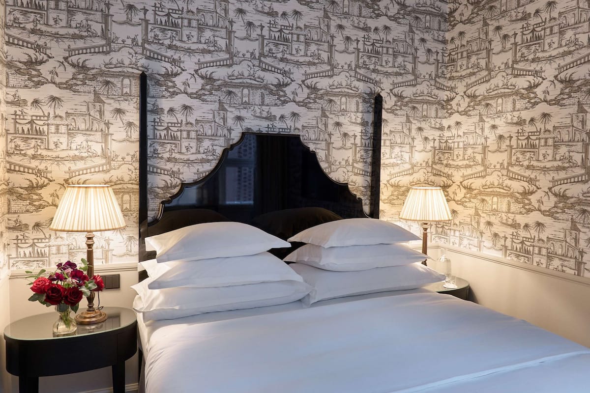 A hotel room with a bed, two bedside tables, and lamps. The walls have a detailed wallpaper design.