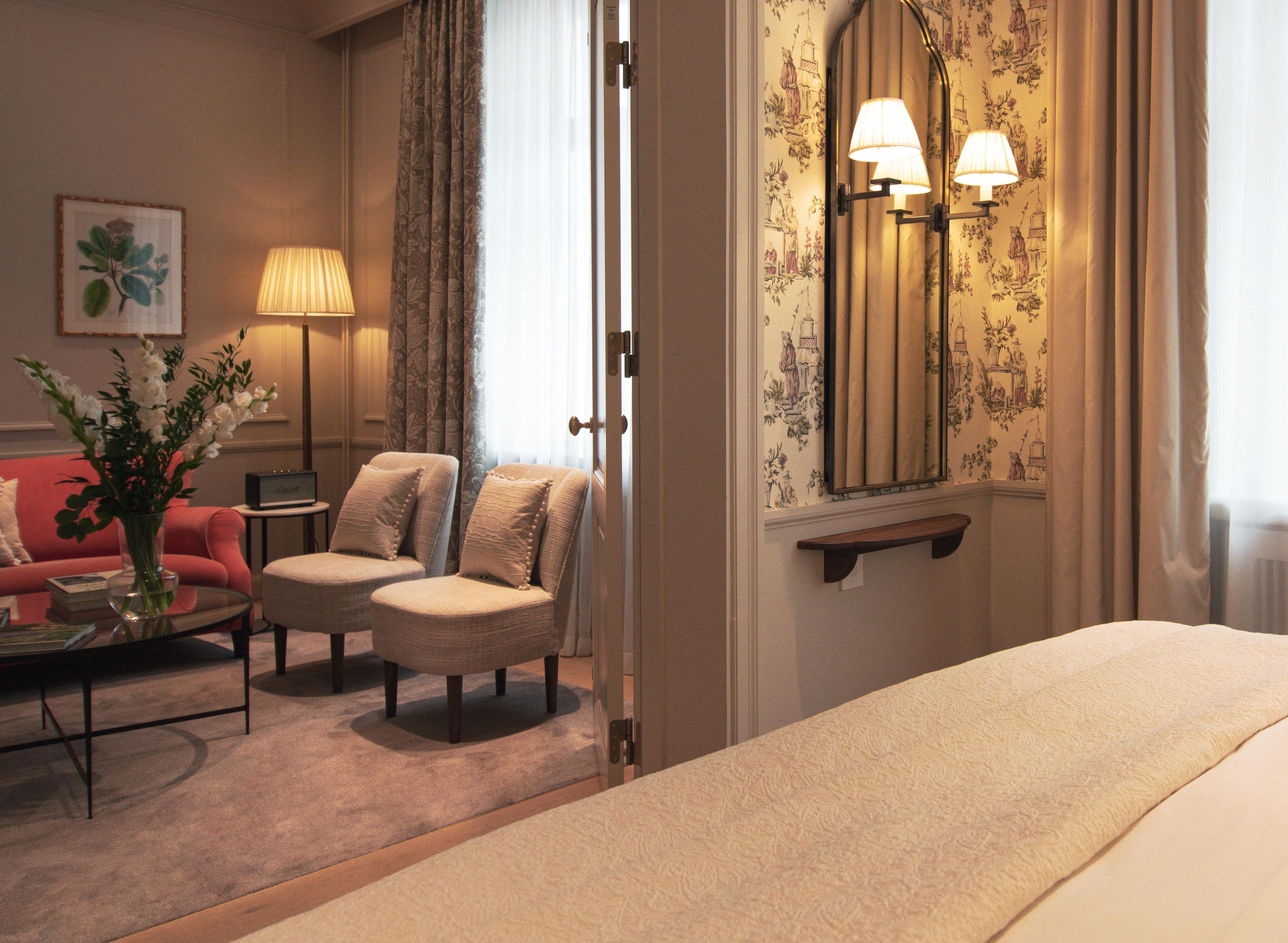 Une chambre d’hôtel avec un lit, deux fauteuils, une petite table avec une lampe, un vase de fleurs et un tableau au mur.