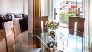 A modern living room with a glass dining table, a sofa, and a view of the city through large windows. - Parque 5 Apart Hotel (Cusco)
